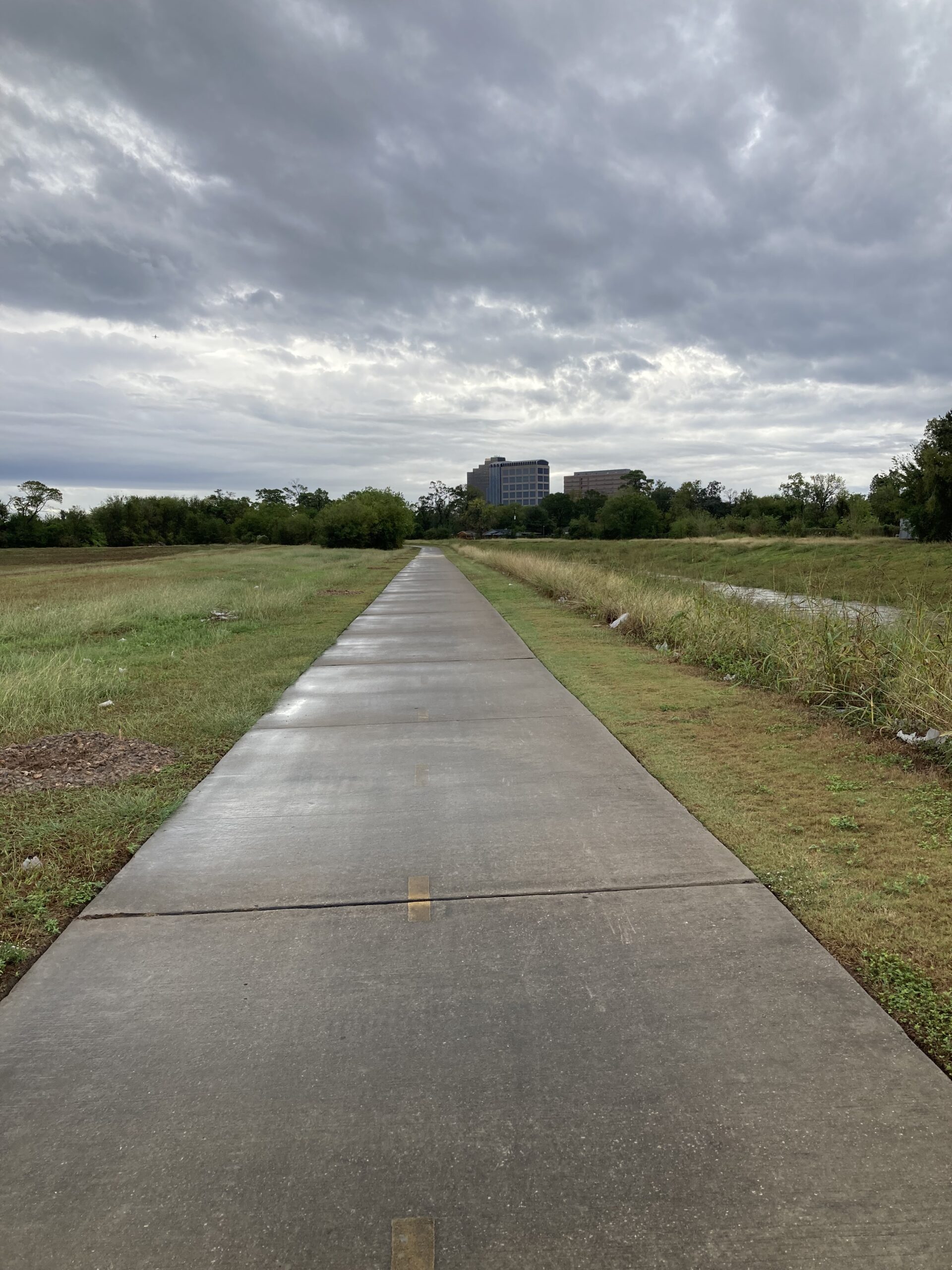 A concrete trail along Greens Bayou on a cloudy day, bordered by grass and sparse vegetation, with office buildings visible in the distance