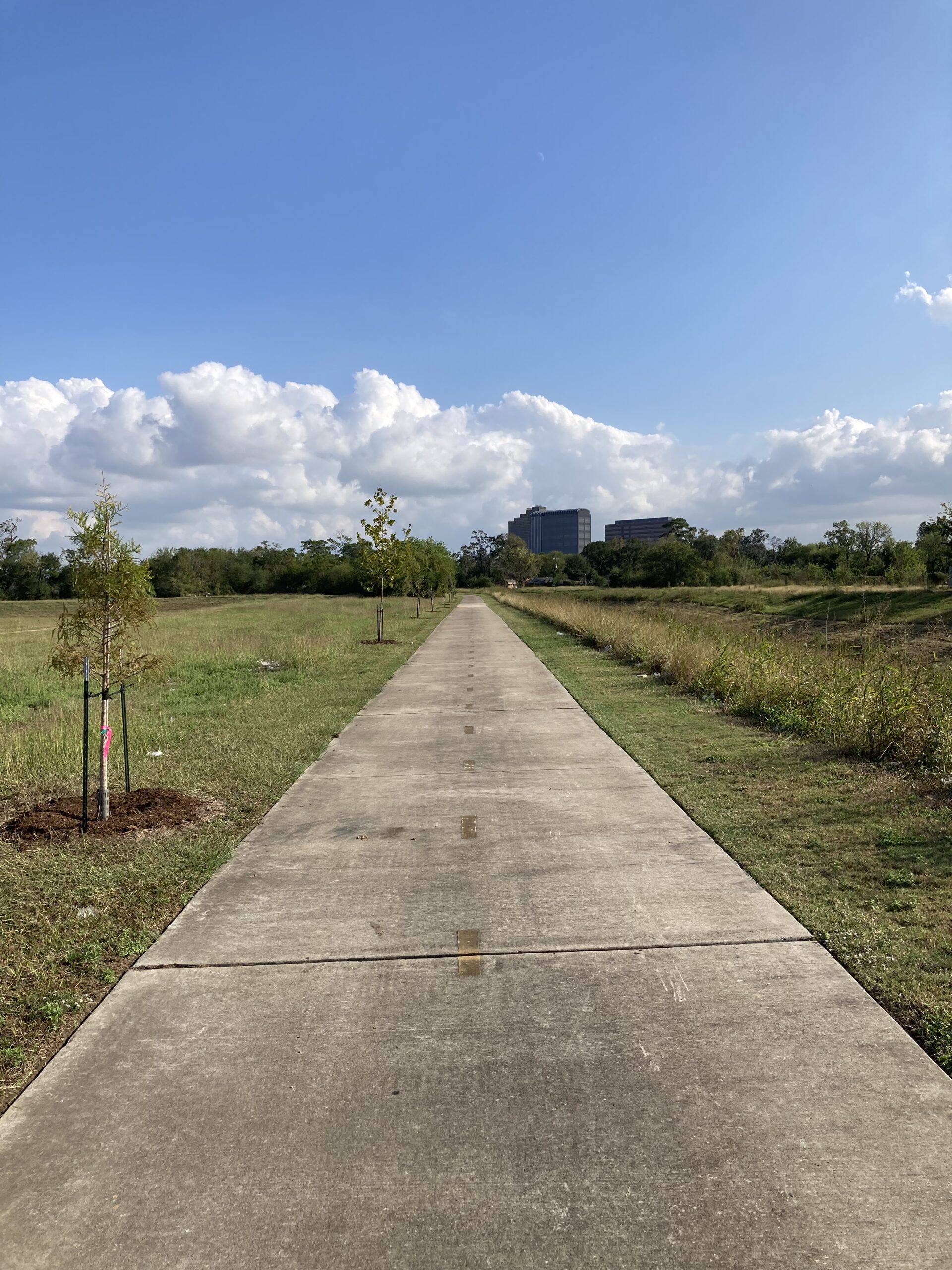 A straight, paved trail along Greens Bayou under overcast skies, with grassy fields and sparse vegetation on both sides of the path, leading toward a distant cluster of buildings