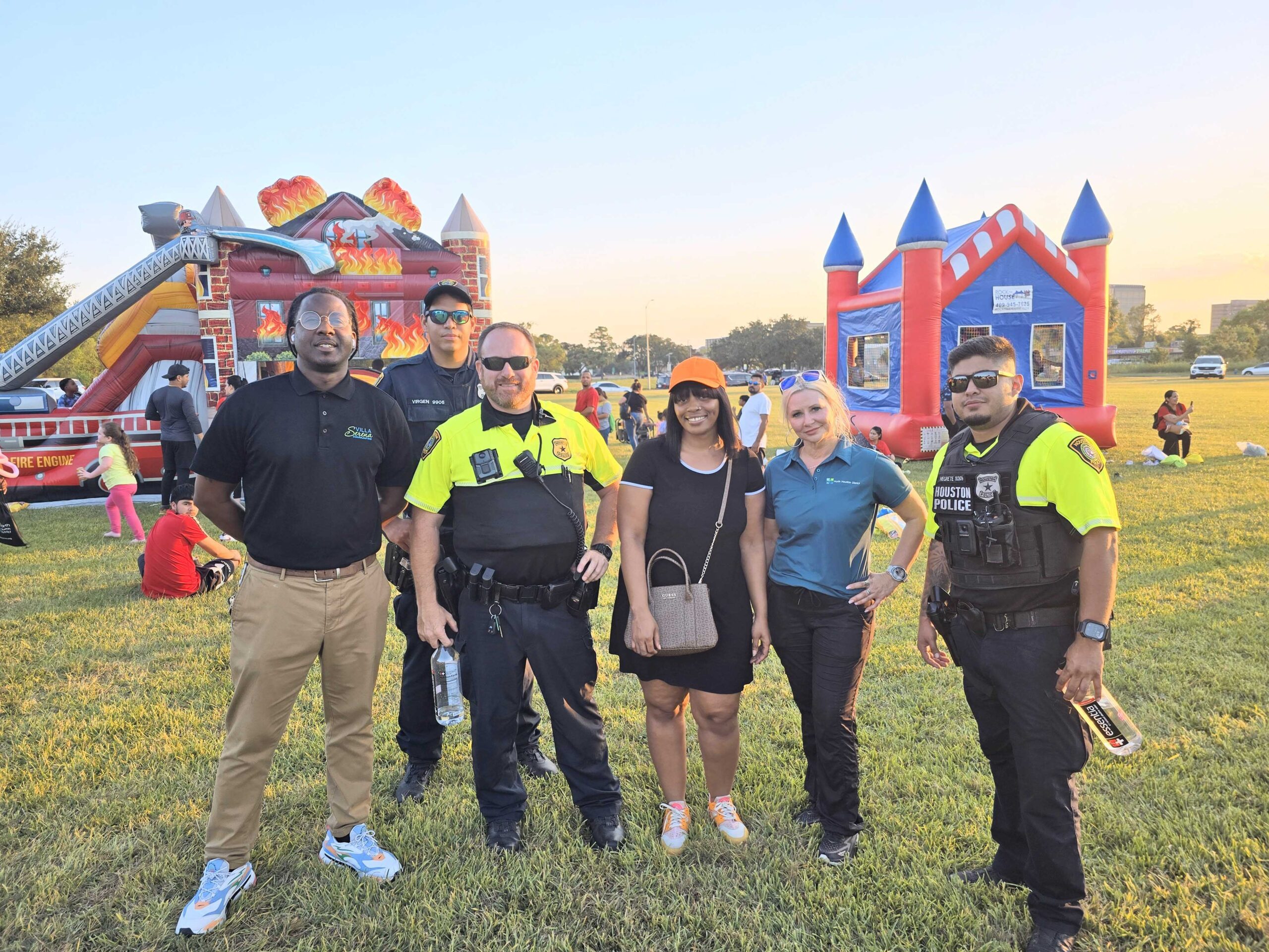 A group photo featuring two police officers, a woman in an orange hat, and two other individuals, standing in front of inflatable bounce houses shaped like a fire truck and a castle. The grassy area is busy with event attendees enjoying the activities in the background.