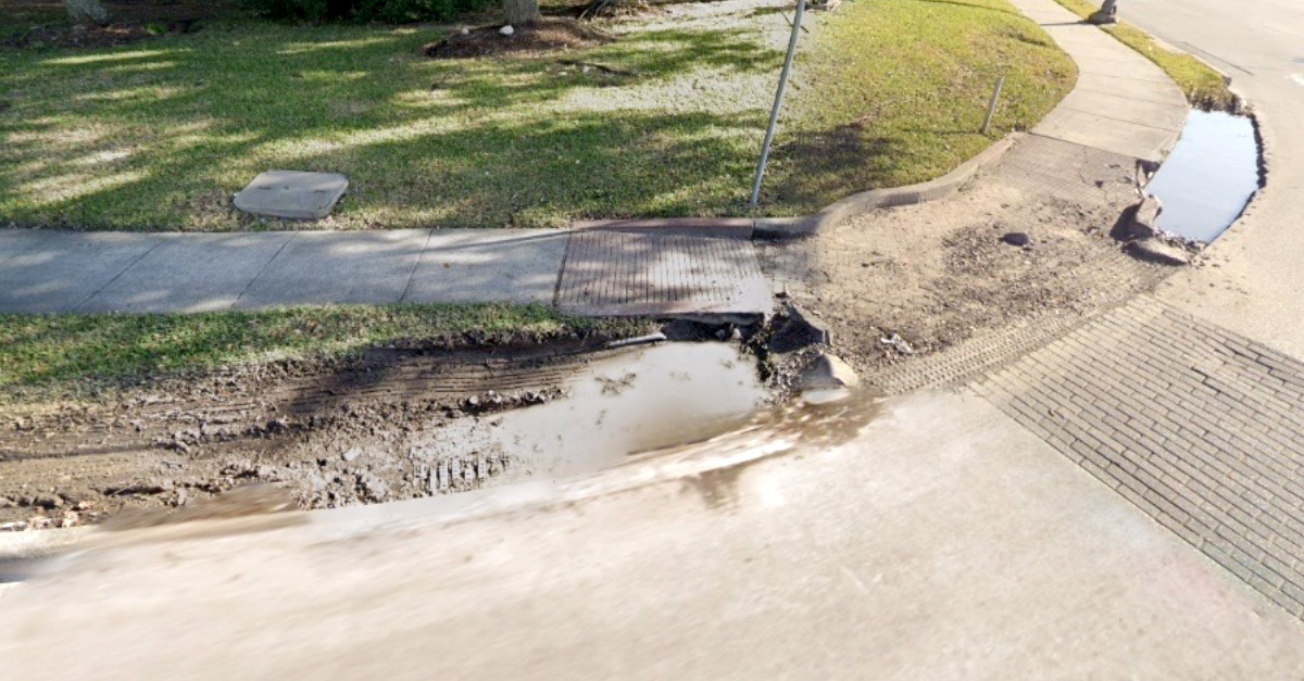 A damaged curb and sidewalk area shows erosion, standing water, and broken pavement near a roadway, illustrating conditions prior to infrastructure improvements.