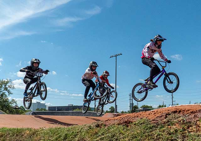 Bike Park 4 Several BMX riders wearing helmets ride closely together over dirt jumps on a BMX track, captured midair against a blue sky during an outdoor cycling event.