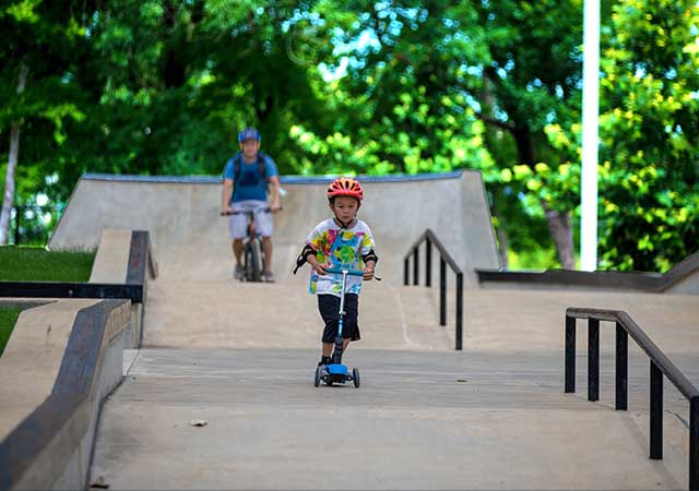 A young child wearing a helmet rides a scooter down a concrete skate park ramp, with an adult on a bicycle following behind among rails and ramps.