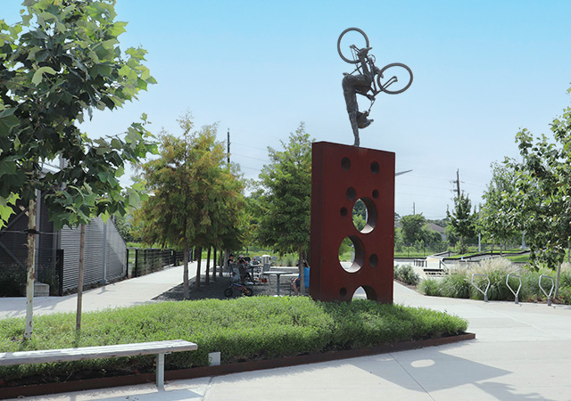 Large BMX-themed sculpture featuring a bicycle rider mounted on a tall red structure overlooking landscaped pathways and park features.