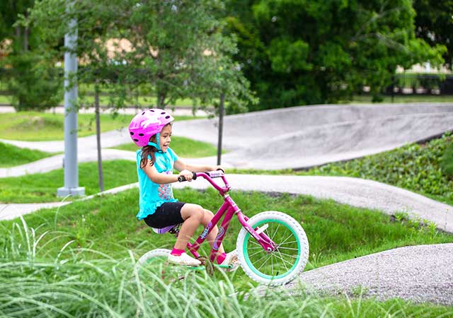 A young child wearing a pink helmet rides a bicycle along a curving paved trail through landscaped greenery at a bike park on a sunny day.