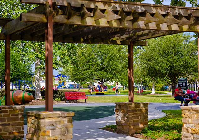 A shaded pergola frames a paved park walkway at Buckboard Park, with red benches, trees, and a playground visible in the background on a sunny day.