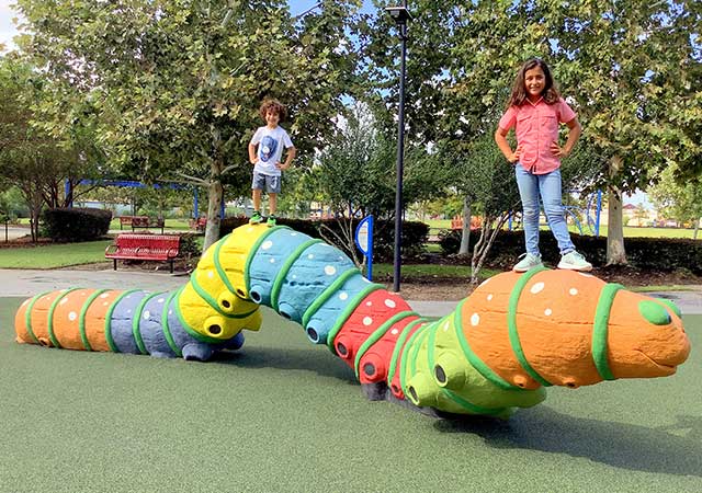 Two children stand and balance on a large, colorful caterpillar-shaped playground sculpture at Buckboard Park, with benches and trees visible behind them.
