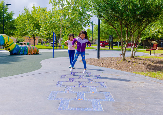Buckboard Park Hopscotch Two children play hopscotch on a mosaic-style pathway feature at Buckboard Park near playground equipment and shaded walking trails.
