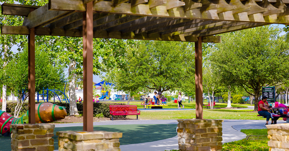 Buckboard Park Newsroom Graphic A shaded pergola frames a view of Buckboard Park, showing playground equipment, benches, walking paths, and families enjoying the park beneath a canopy of trees.