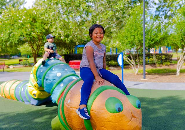 A child smiles while sitting atop a colorful caterpillar-shaped playground sculpture at Buckboard Park, with another child seated further back and trees surrounding the play area.
