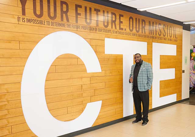 A man in a green plaid jacket stands in front of a large CTE sign with the quote Your Future. Our Mission.