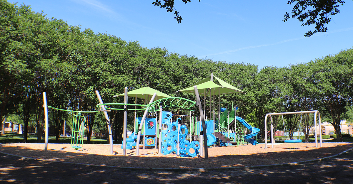 Wide view of the playground at City View Park featuring slides, climbing structures, swings, shaded canopies, and mulch surfacing surrounded by mature trees and greenspace.
