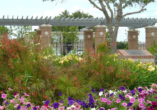 Colorful flower beds fill the foreground at City View Park, with a pergola supported by brick columns and mature trees visible behind the landscaped area.