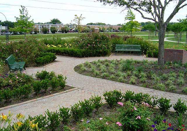 A brick-paved walkway curves through a landscaped garden at City View Park, featuring benches, shrubs, flowering plants, and open green space beyond.