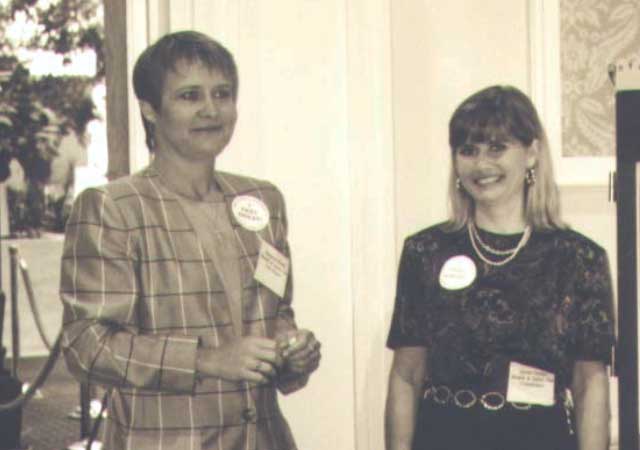 A black-and-white photo of two women standing together at an event. One woman on the left is wearing a plaid jacket, and the woman on the right is wearing a black blouse and skirt. Both women are smiling, and they are wearing event badges.