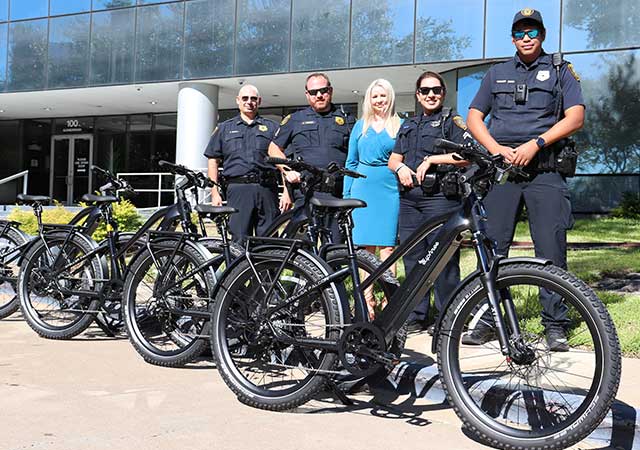 Five uniformed officers and a woman in a blue dress stand behind a line of black e-bikes in front of a glass building. They smile for the camera on a sunny day.