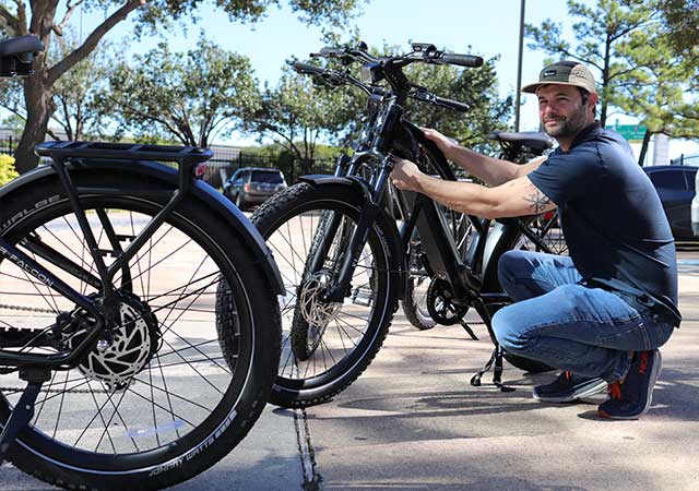 A man in a baseball cap crouches near a black e-bike, inspecting it. Additional e-bikes and a parking lot are visible in the background under a clear blue sky.