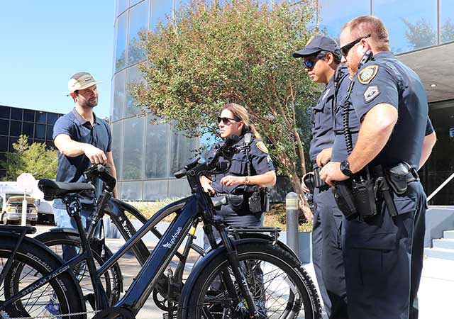 A man explains features of black e-bikes to three uniformed officers in a group discussion. They stand outside near a glass building and trees on a sunny day.