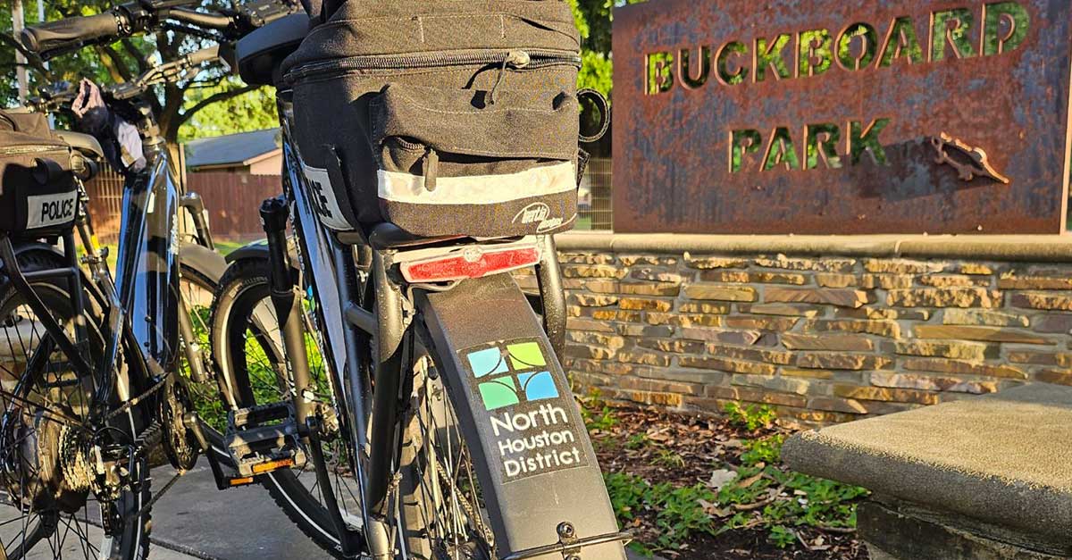 Two black North Houston District-branded police e-bikes are parked near the Buckboard Park sign, with greenery and a walking path in the background.