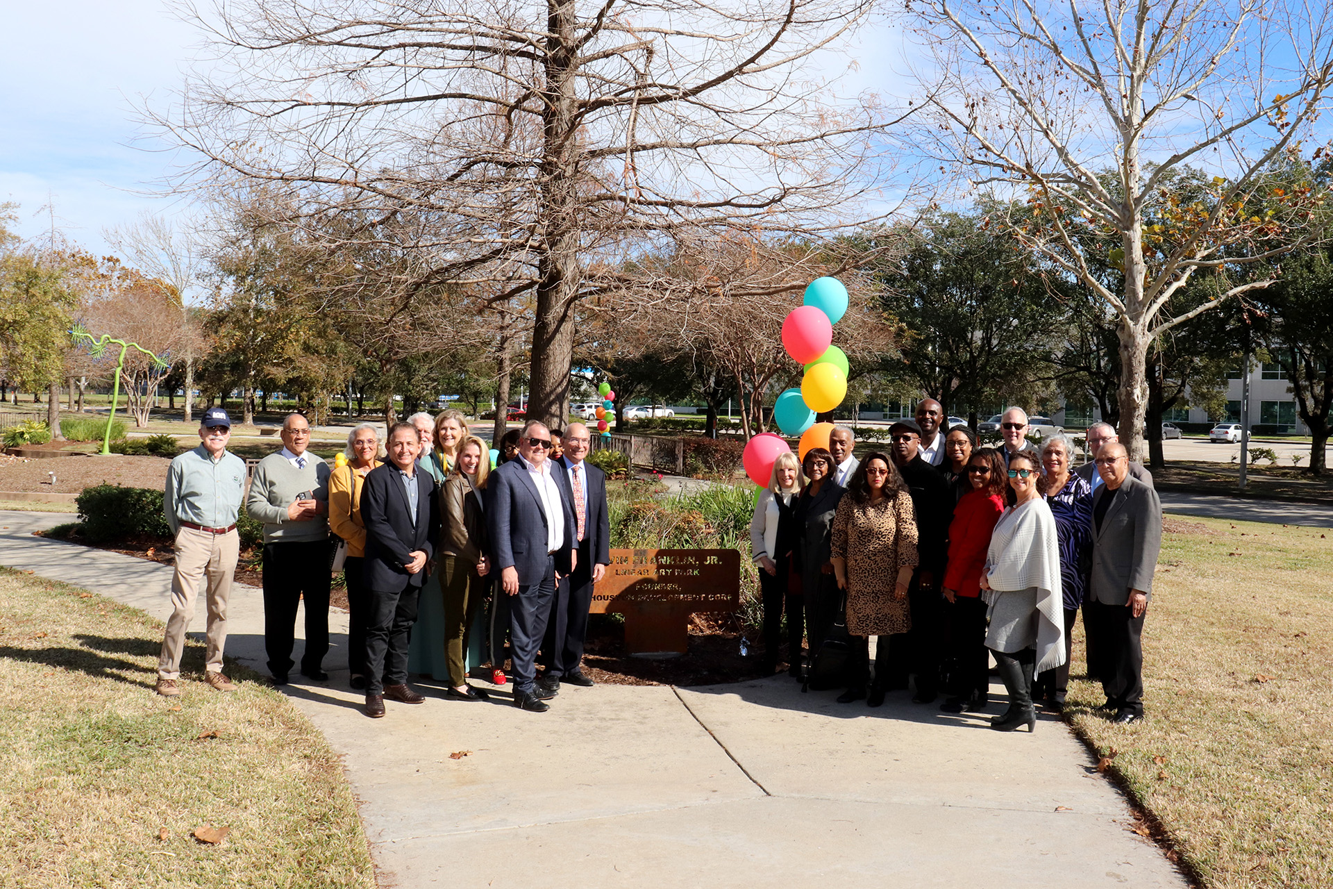 A large group of attendees, dressed in professional and semi-formal attire, pose together in front of the park plaque with colorful balloons.