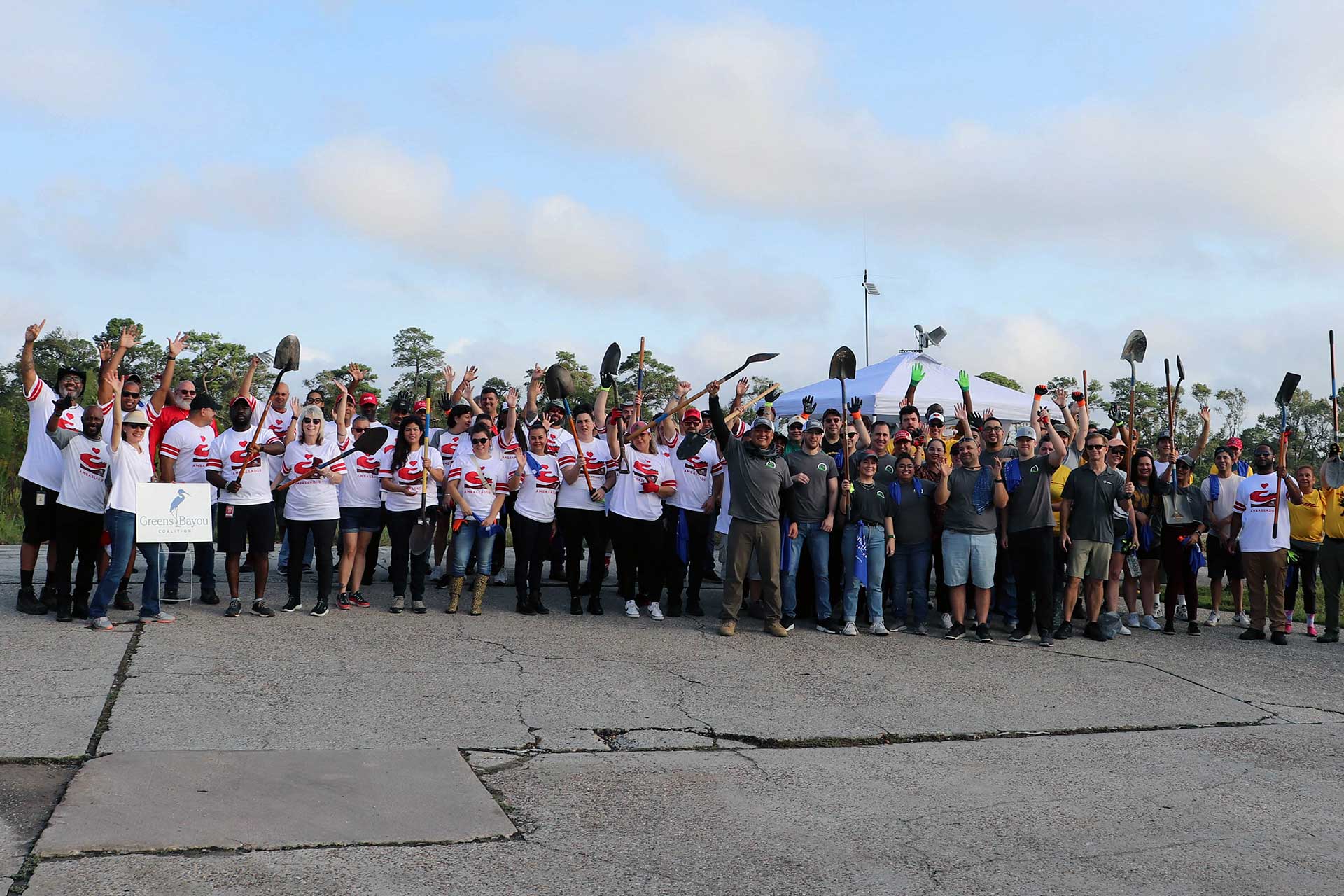 A large group of volunteers from the previous photo raises their hands and tools, such as shovels and gloves, in a celebratory pose. The scene includes a bright sky and grassy surroundings.