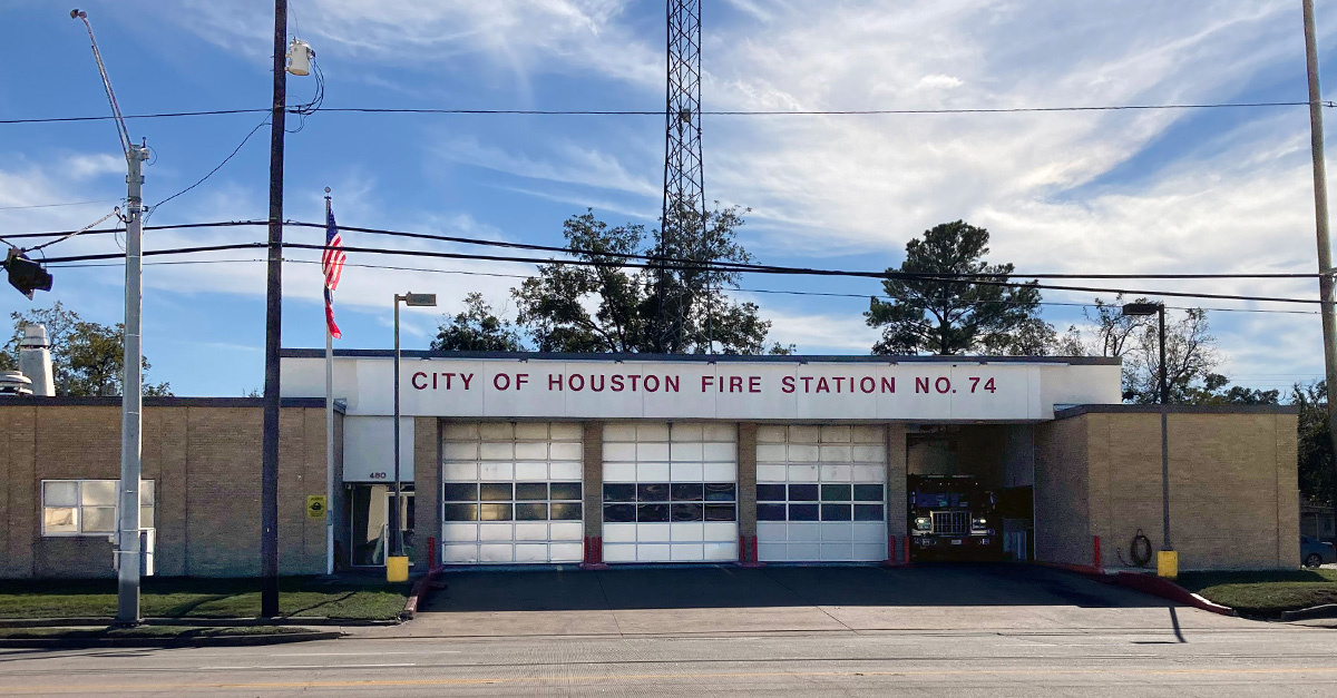 The exterior of City of Houston Fire Station No. 74 shows a brick fire station with multiple bay doors, an American flag, and a fire engine visible inside one open bay along a city street.