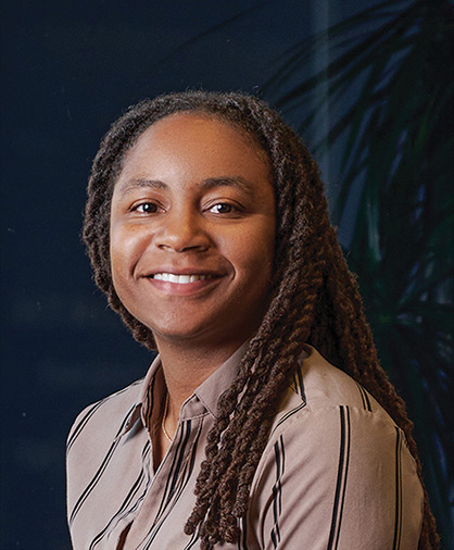 Headshot of Hershiira Boone-Rodgers smiling, wearing a striped blouse, in front of a dark blue background with a leafy plant in soft focus.