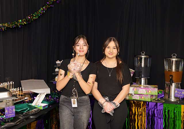 Two students wearing gloves stand in front of a Mardi Gras-themed hospitality setup with food and drink tables.