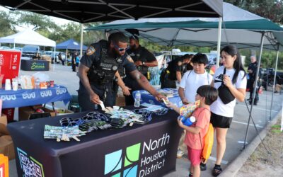 Two children, a boy and a girl, are receiving sunglasses and small items from a police officer at a booth. The officer is handing an item to one of the children while another child watches. The table is filled with giveaways, and two police cars are visible in the background as part of the National Night Out event.