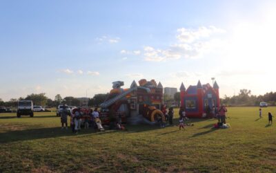 A wide shot of a grassy field with two large inflatable bounce houses, one shaped like a fire truck with a slide, and the other a traditional castle bouncer. Several children and adults are gathered around the inflatables, enjoying the sunny day. The sky is clear with a few scattered clouds, and there are cars parked in the distance.
