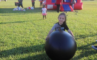 A smiling young girl in a gray shirt with the words "Char Rush" on it is playing with a large black ball on a grassy field. Behind her, there are other children, and an inflatable bounce house in red and blue is visible in the background.