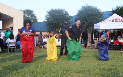 A lively sack race with four participants. A child in a yellow sack, a law enforcement officer in a green sack, and two others in red and blue sacks, respectively, are all hopping on a grassy area, with spectators and more event activities visible behind them.