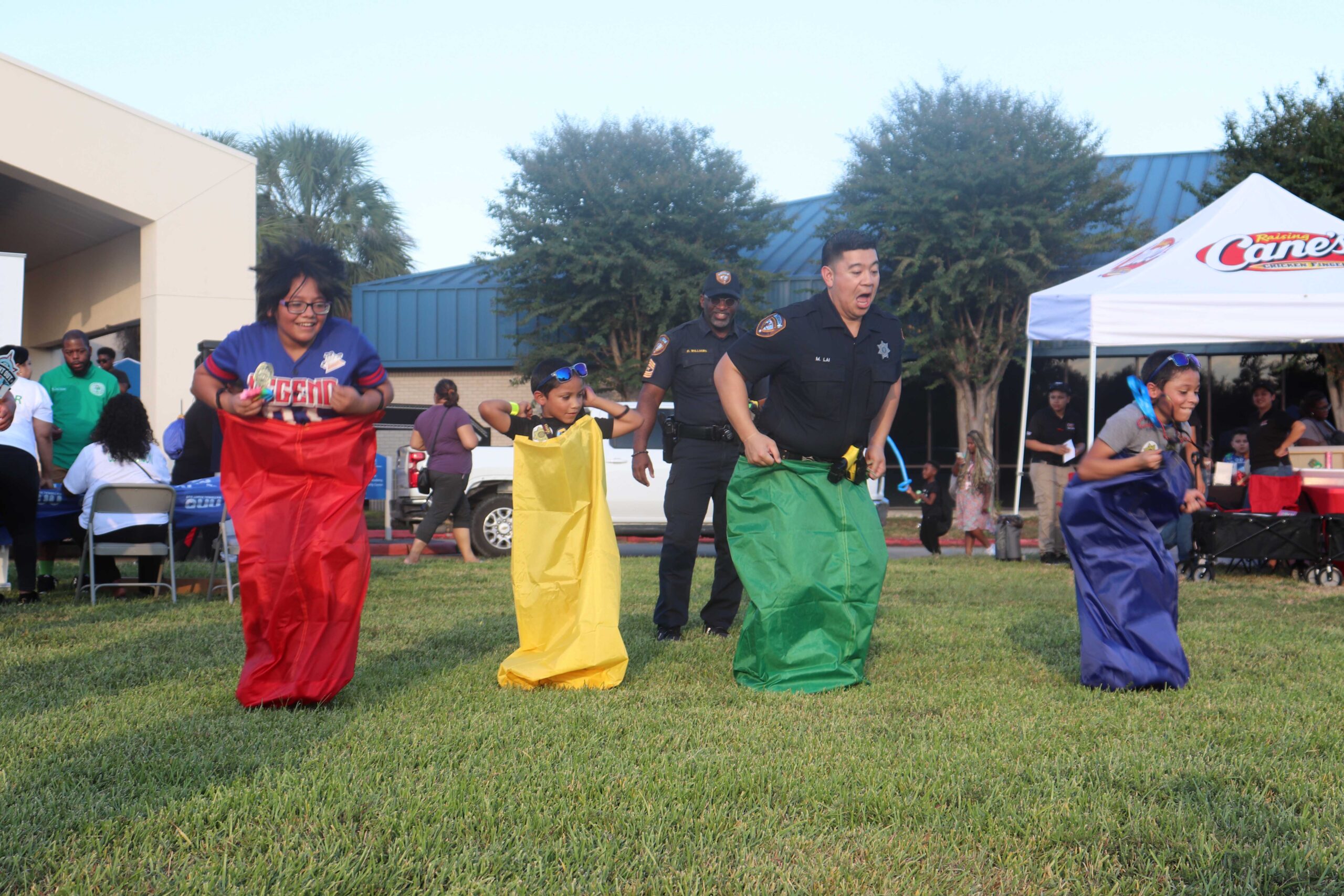 A lively sack race with four participants. A child in a yellow sack, a law enforcement officer in a green sack, and two others in red and blue sacks, respectively, are all hopping on a grassy area, with spectators and more event activities visible behind them.