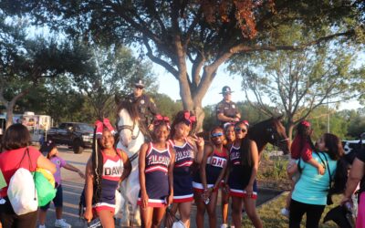 A group of seven cheerleaders, wearing navy blue, red, and white uniforms with "DHS" and "Falcons" on them, are posing with two mounted police officers. The officers are sitting on their horses, and the cheerleaders are standing and smiling in front of them. Other people are visible in the background, along with trees and a parked car.