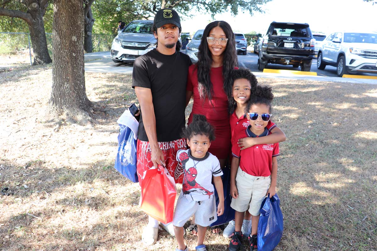 A family of five smiles at the camera, standing together outdoors on a grassy area. The mother, dressed in a red dress, and father, in a black T-shirt and hat, stand with their three children, two of whom wear matching red shirts, while the youngest child wears a Spider-Man T-shirt. They hold blue and red goodie bags from the event.