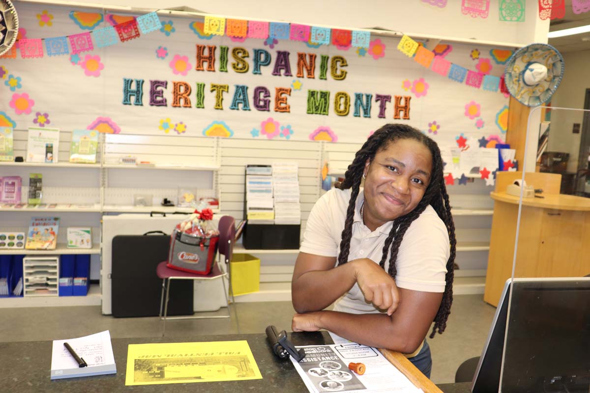 A smiling woman leans on a counter in front of a colorful "Hispanic Heritage Month" display at a library. The banner is decorated with vibrant paper flowers and banners. Informational flyers and books are visible in the background.