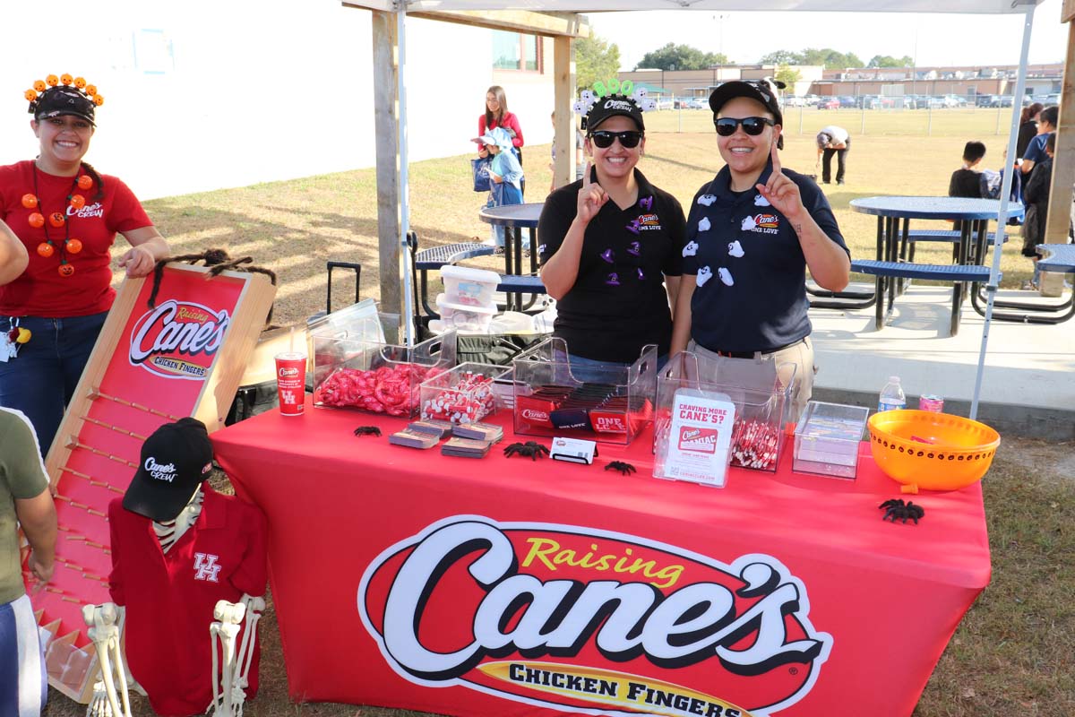 Three Raising Cane's employees stand behind a booth decorated with Halloween items, including a game board and various giveaways. The table is covered with a red Raising Cane's tablecloth, and the employees, dressed in themed attire with headbands, smile at the camera.