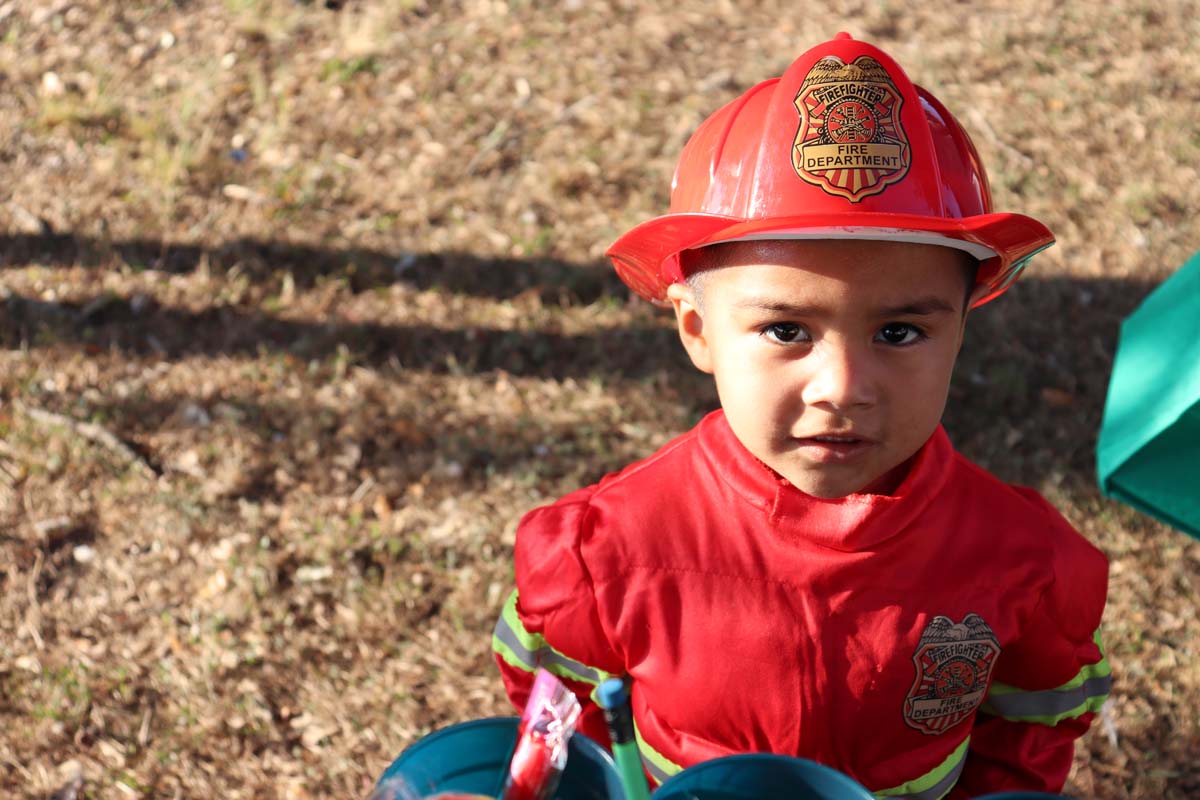 Young Boy in Firefighter Costume: A young boy dressed in a red firefighter costume with a plastic firefighter hat looks directly at the camera.