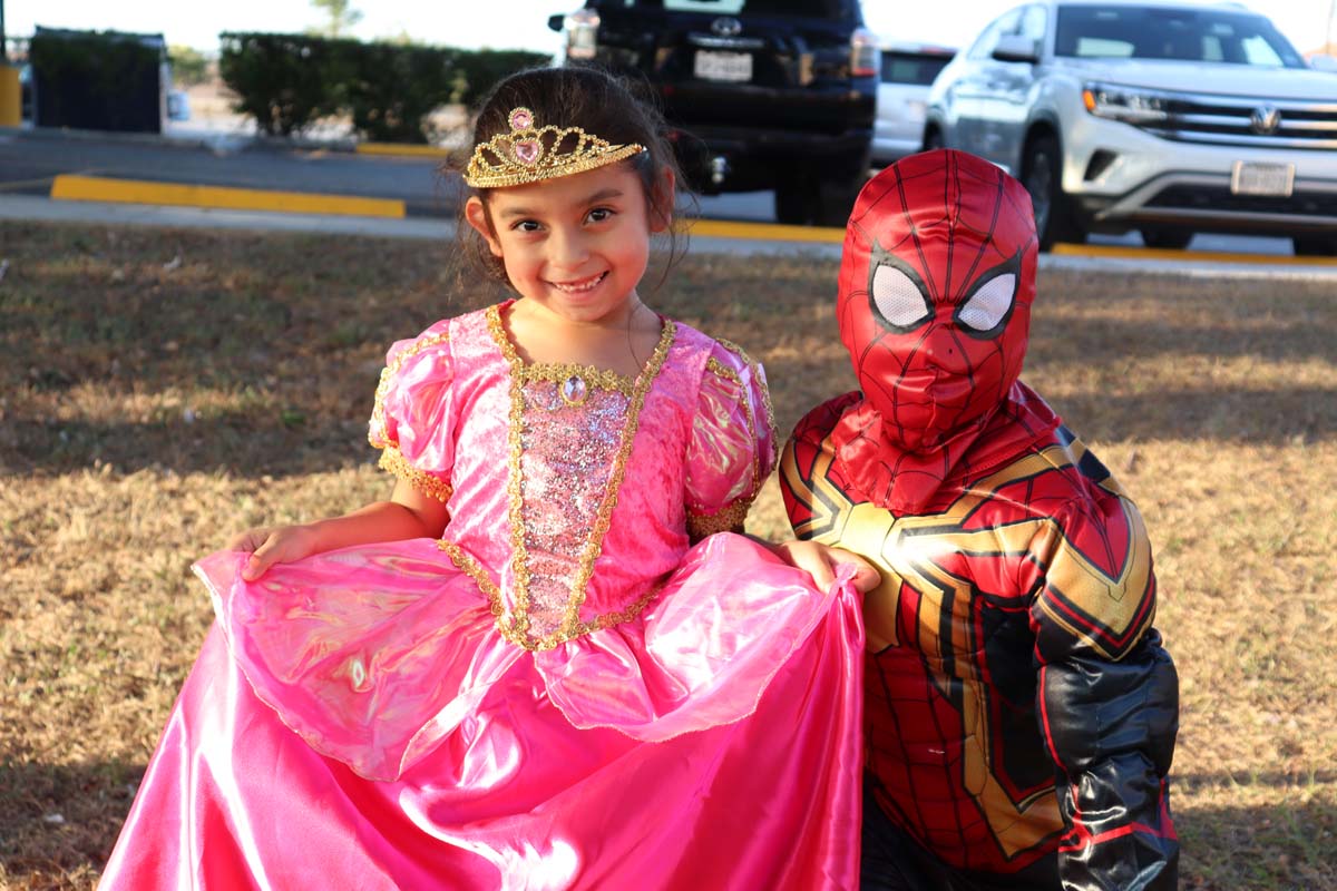 A young girl dressed in a vibrant pink princess gown and tiara smiles at the camera while holding out her skirt. Next to her, a boy dressed as Spider-Man poses, both enjoying the Halloween event outdoors.