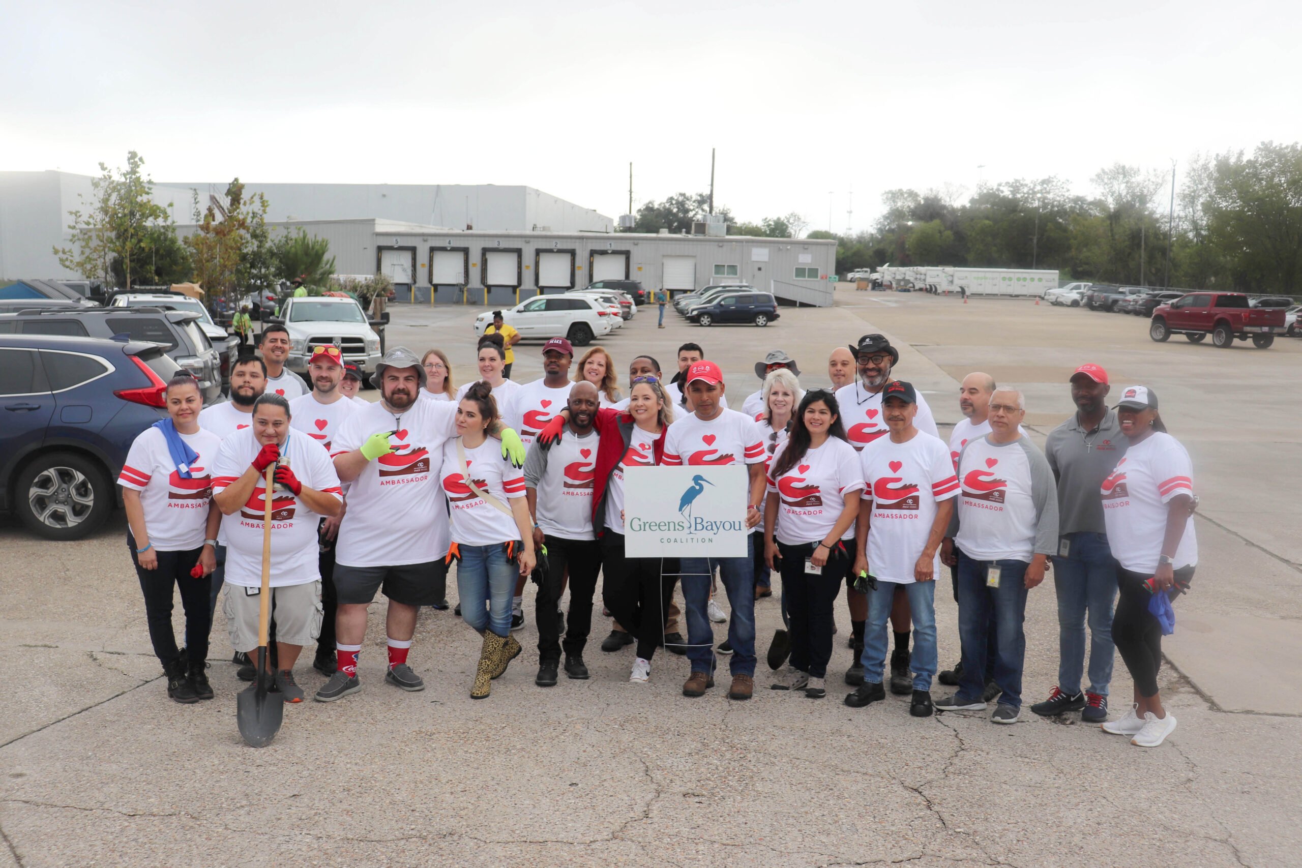 A large group of volunteers wearing matching white t-shirts with red logos pose for a group photo in a parking lot. They stand behind a "Greens Bayou Coalition" sign and are smiling, holding tools like shovels and gloves.