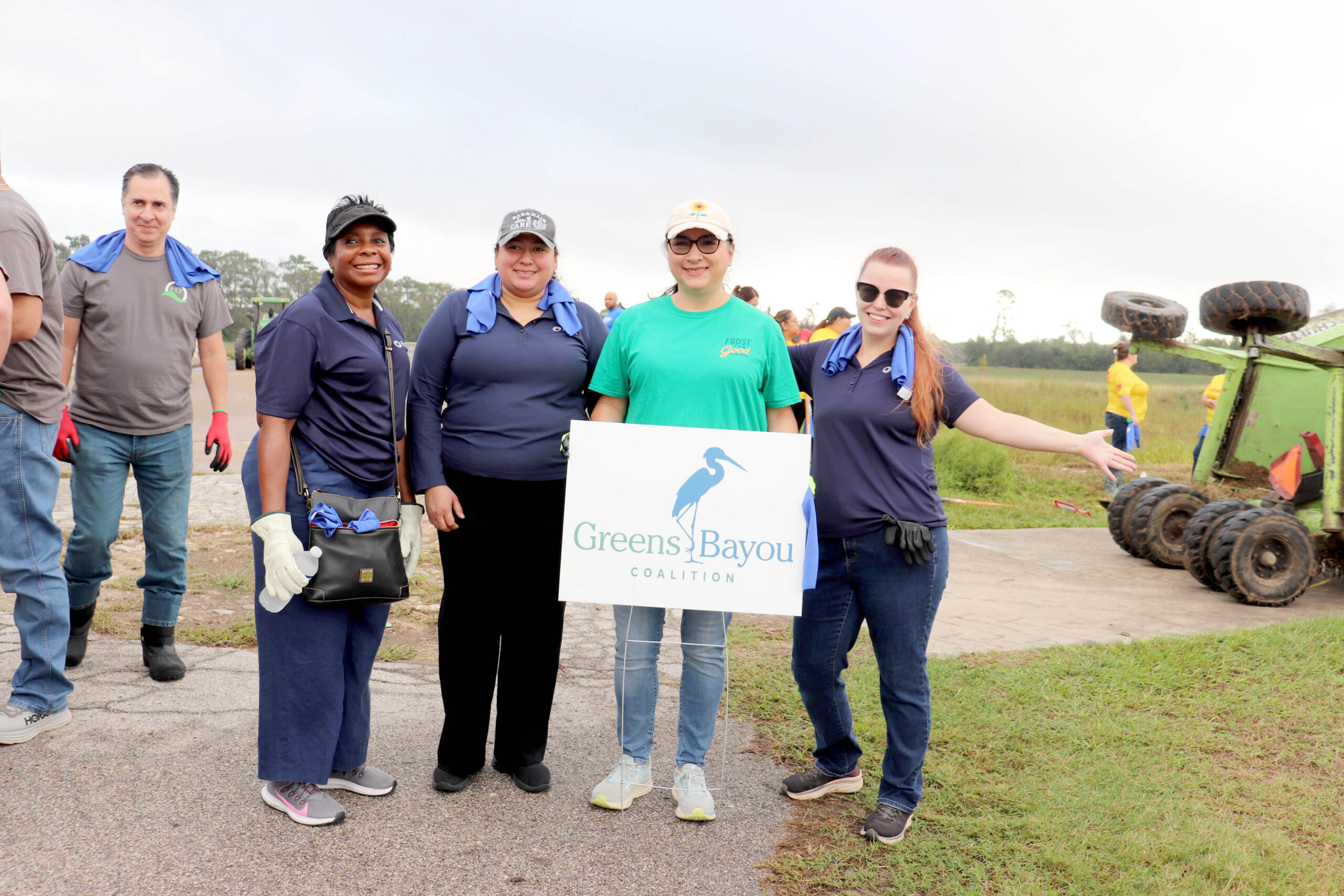 Four volunteers stand together holding a "Greens Bayou Coalition" sign. They are outdoors on a grassy trail, wearing navy and green shirts, and smiling. A vehicle with large wheels is in the background.