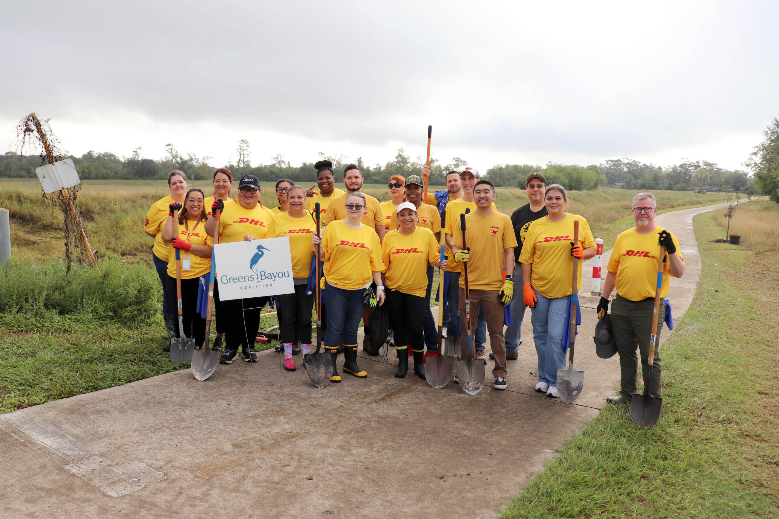 A group of volunteers in yellow shirts with "DHL" logos poses on a paved trail surrounded by grass. They hold shovels and stand behind a "Greens Bayou Coalition" sign, smiling under a cloudy sky.