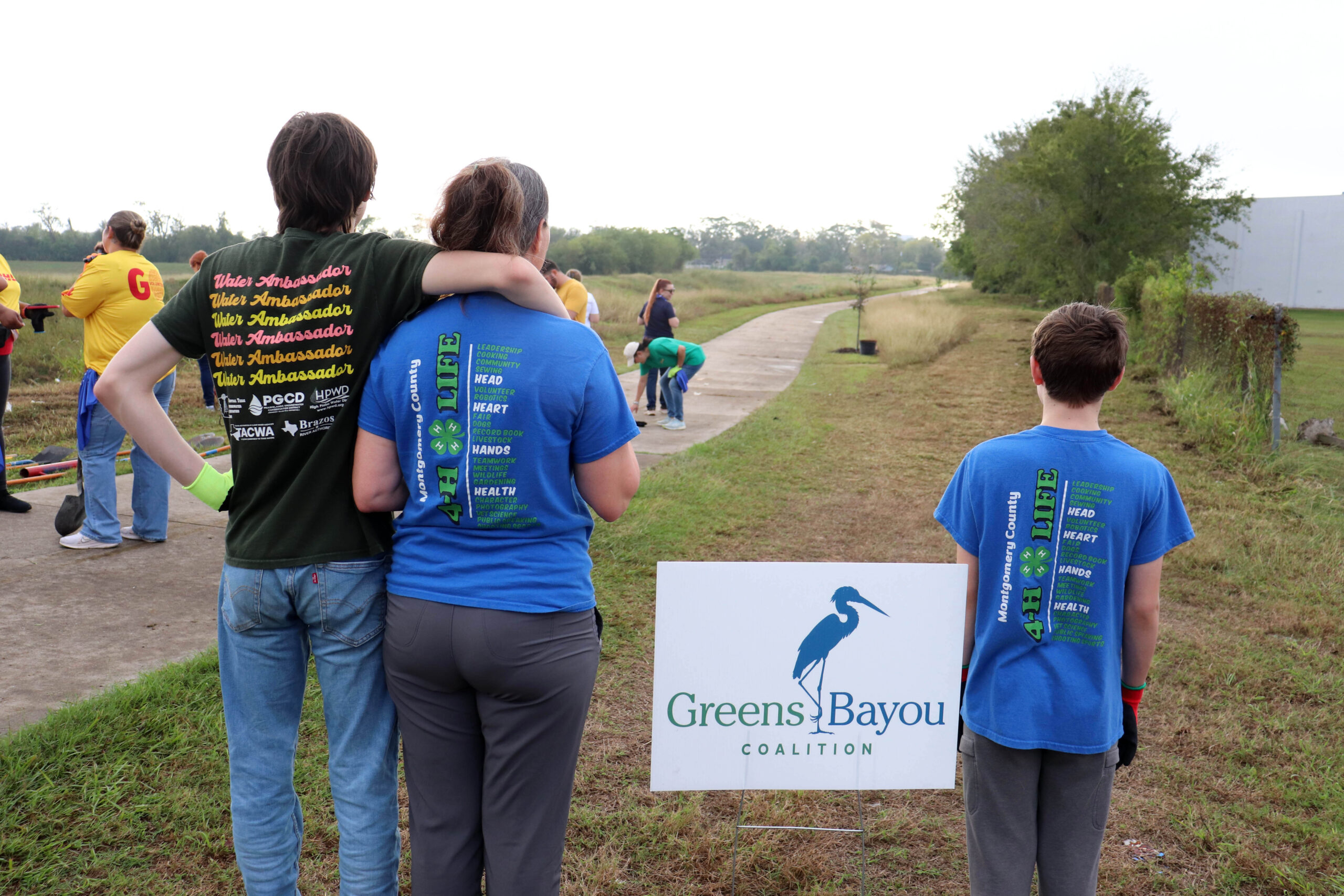 Two adults and a child in blue shirts face away from the camera, standing next to a "Greens Bayou Coalition" sign. They are on a grassy trail, embracing and looking toward a natural landscape.