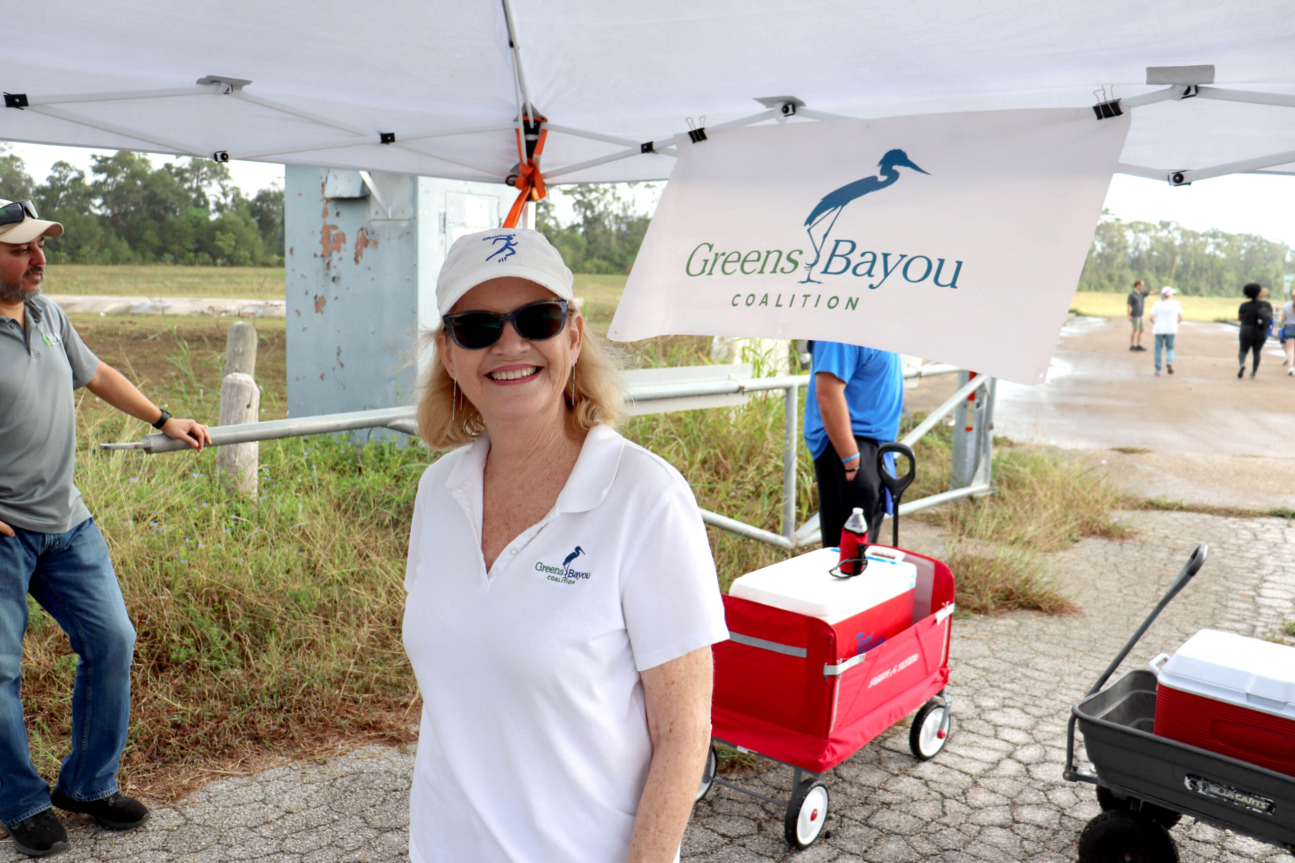 A woman wearing a white "Greens Bayou Coalition" polo shirt and cap smiles under a canopy with a "Greens Bayou Coalition" banner. Red and gray wagons are visible nearby, along with grassy surroundings and other volunteers in the background.