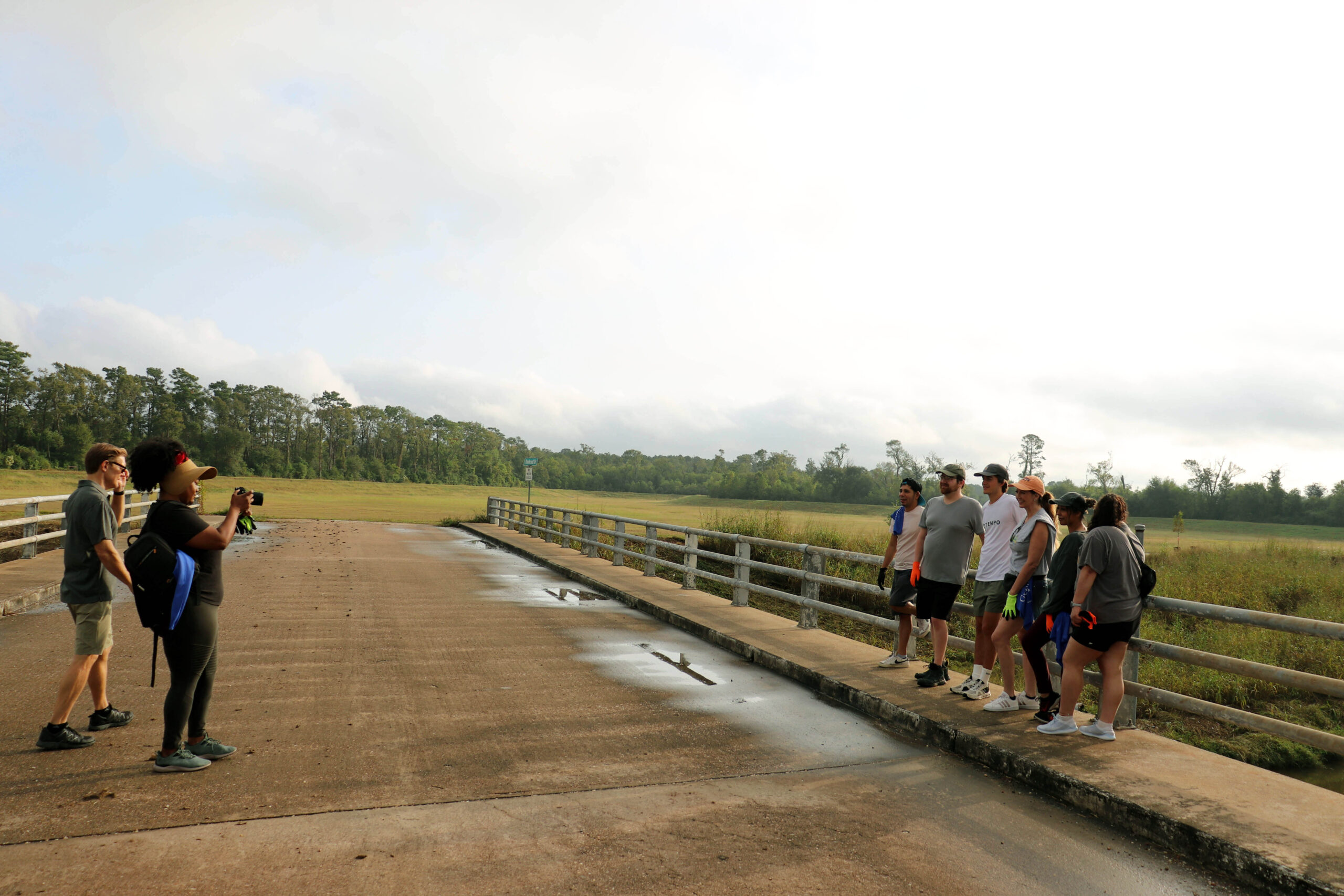 Volunteers are gathered on a bridge in a natural setting. A photographer captures a group of people lined up along the railing, with trees and open fields in the background under a cloudy sky.