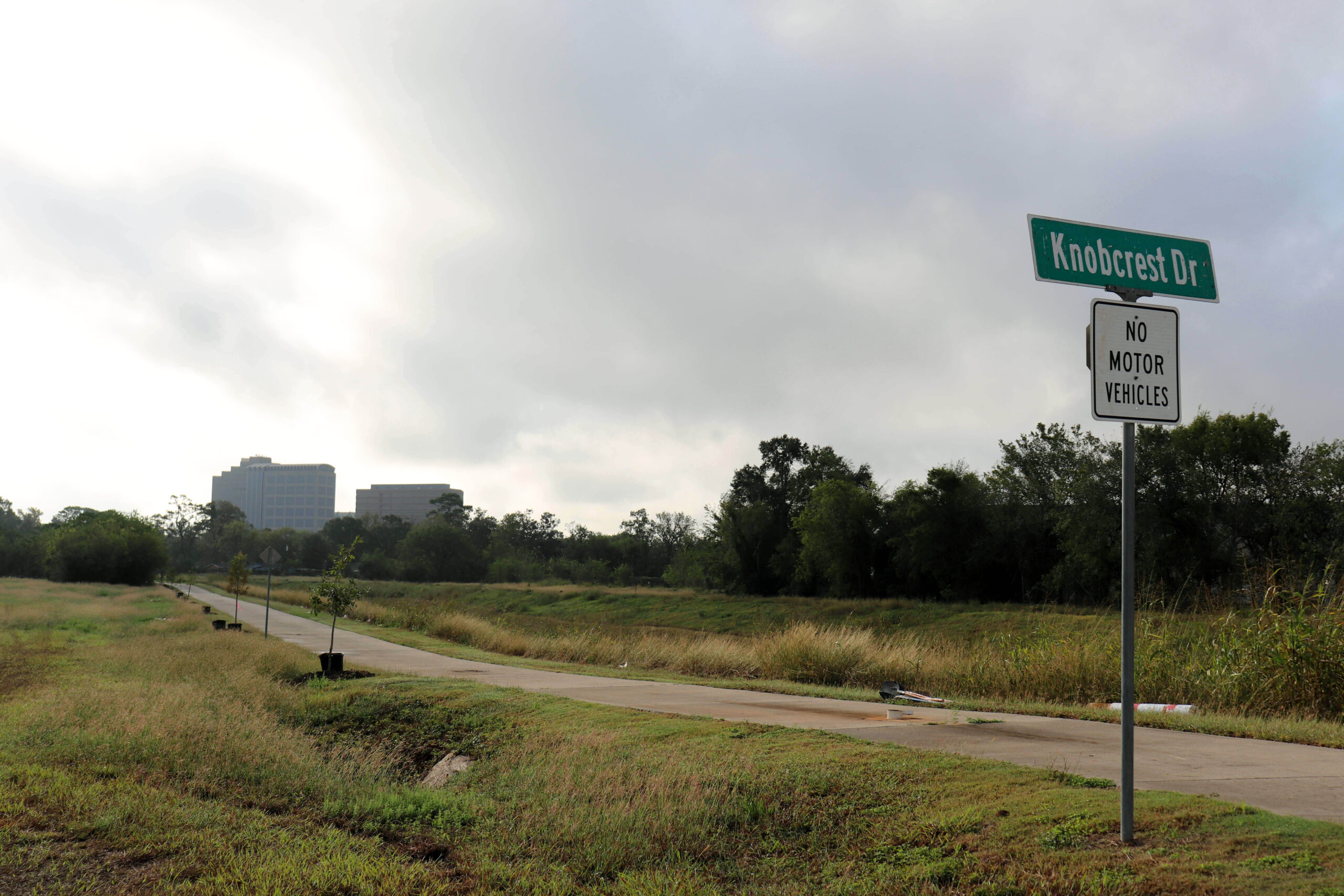 A paved trail in a grassy area is shown with a "Knobcrest Dr" sign and a "No Motor Vehicles" sign in the foreground. The path is lined with small planted trees, and buildings are visible in the distance.