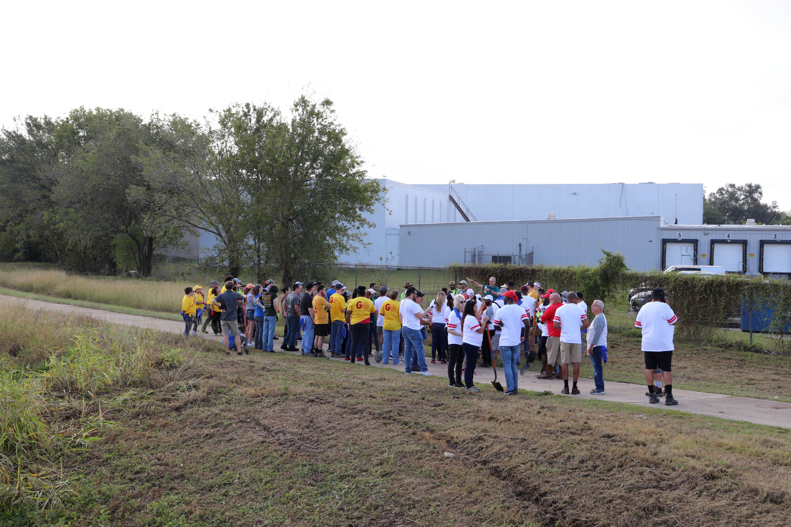 Volunteers gather in a larger group near a paved path surrounded by grass and trees. Many are wearing matching shirts, and the background includes a building with loading docks.