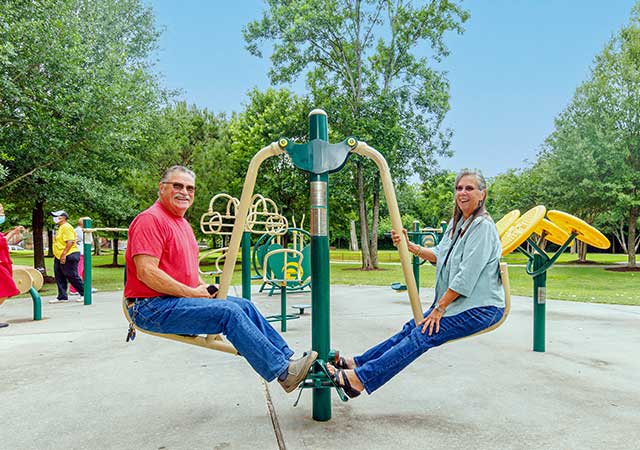 Two adults sit facing each other on an outdoor fitness machine at Ida Gaye Gardens, smiling as they exercise on a concrete pad surrounded by trees and open green space.