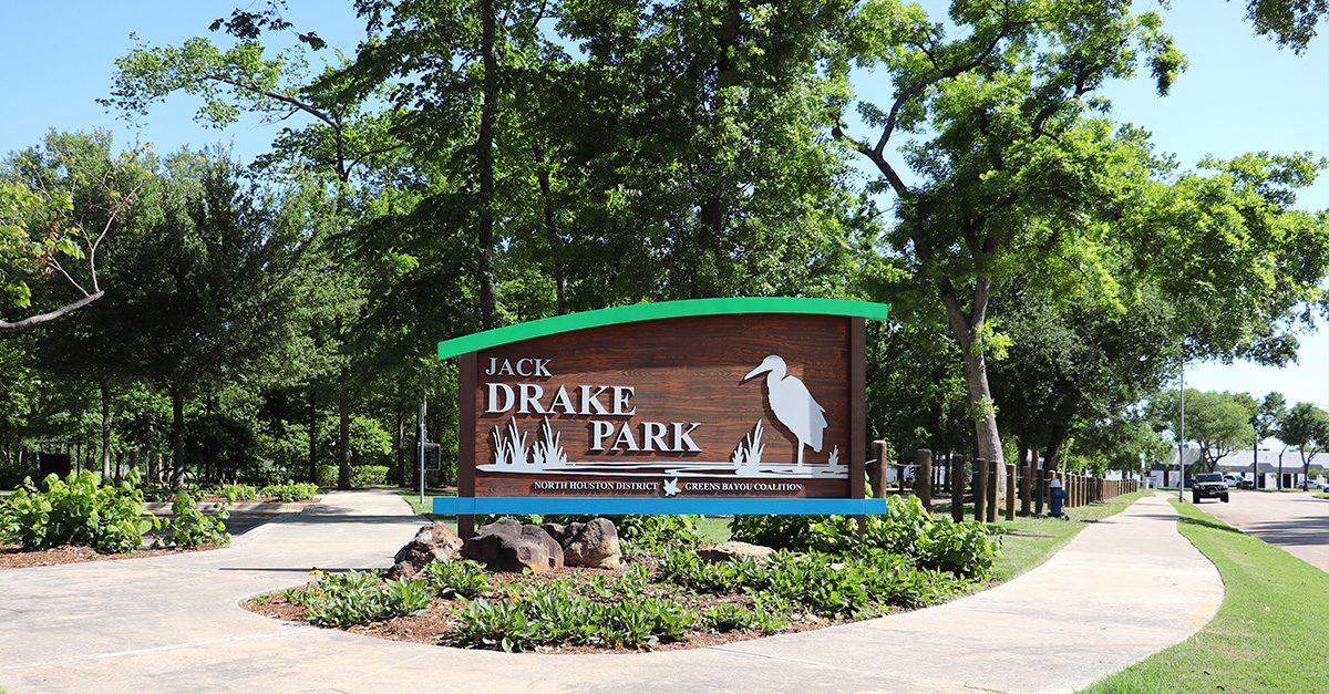 Entrance sign for Jack Drake Park features a wooden design with a white heron silhouette and green trim, surrounded by trees, landscaped plants, and a walking path under a clear blue sky.