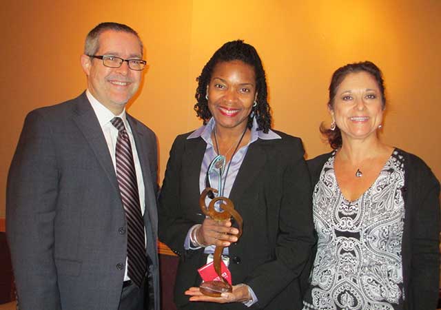 Karen Marshall, holding a metal award, is pictured with two colleagues dressed in business attire. The trio smiles warmly in front of a softly lit yellow wall.