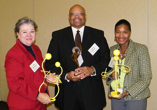 Karen Marshall stands on the right, holding a yellow sculpture award, smiling alongside two colleagues who also hold awards. The group is dressed in business attire and poses in front of a beige wall.